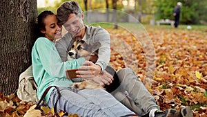 Loving Couple Man And Woman sitting with Welsh Corgi Dog In City Park hugging and making selfie on smartphone