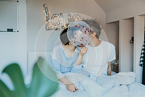 Loving couple in pajamas kissing behind a balloon with red hearts inside in bed with word Love on the wall background