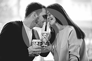 Loving couple is siting face to face on the background of window and holding cups in their hands. Black and white photo