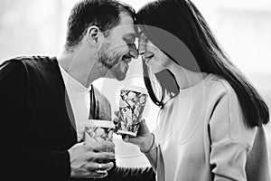 Loving couple is siting face to face on the background of window and holding cups in their hands. Black and white photo