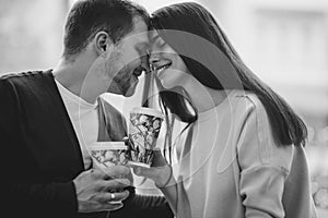 Loving couple is siting face to face on the background of window and holding cups in their hands. Black and white photo