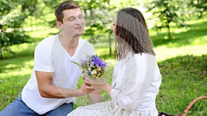 Loving couple sits on blanket in park during picnic, man gives woman bouquet of wild flowers, compliments woman, and