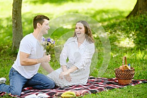 Loving couple sits on blanket in park during picnic, man gives woman bouquet of wild flowers, compliments woman, and