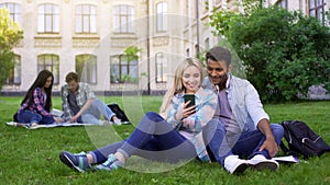 Loving couple of students sitting on lawn and watching video on smartphone