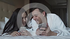 Loving couple in white coats on bed happily share information in their iPhones slow motion stock footage video