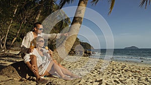 Loving couple in white dress and sunglasses, near palm tree, relax and drinking cocktail. Concept sea beach tropical