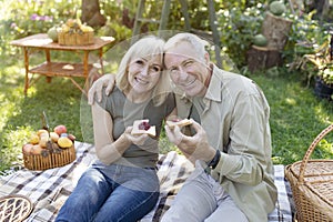 Loving elderly spouses having picnic, sitting on blanket in garden and eating toasts with jam, smiling at camera