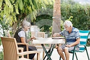 Loving elderly spouses resting in garden and enjoying days, sitting outdoors.Woman reading book, man in front of her