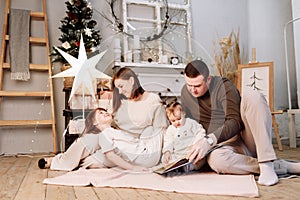 Loving family. Mom, dad with little daughters read interesting book and having fun near decorated fir tree and fireplace