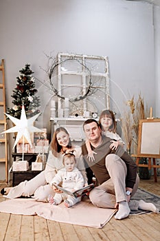 Loving family. Mom, dad with little daughters read interesting book and having fun near decorated fir tree and fireplace