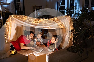 Loving family, mother, father and daughter lying inside self-made hut, tent in room in the evening and reading book