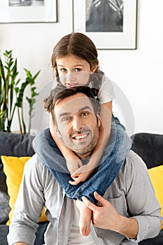 Loving father carrying his little toddler daughter on the shoulders sitting on the sofa in cozy living room, single