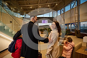 Loving father hugs his son and wife as she smiles, looking over her shoulder, pointing to timetable information panel, checking