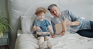 Loving father reading book to child while kid using tablet in bed in apartment