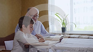 Loving grandfather shows his beloved little grandson a family photo album sitting at a table