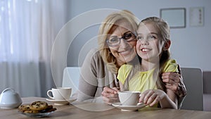 Loving grandma drinking tea with cute granddaughter, tightly hugging her, family