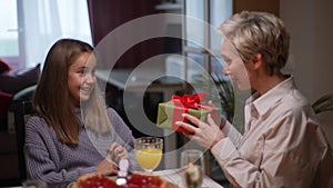 Loving grandmother giving festive box with Christmas present to adorable granddaughter sitting at dinner feast table