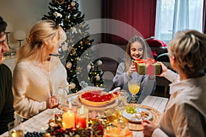 Loving grandmother giving festive box with Christmas present to cheerful granddaughter sitting at dinner feast table