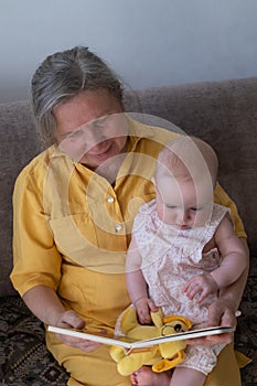 Loving grandmother reading a book to baby girl holding book sitting on sofa.