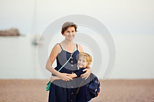 Loving grandson tenderly embracing his joyful mature grandmother while walking on sea shore. Two generations of family