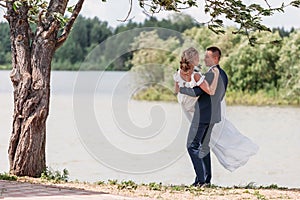 A loving groom circles bride in his arms on the field under tree in nature