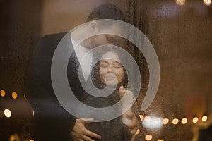 Loving guy hugs and kisses his girlfriend standing behind a wet window with lights. Romantic couple