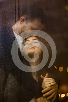 Loving guy hugs and kisses his happy girlfriend standing behind a wet window with lights. Romantic couple