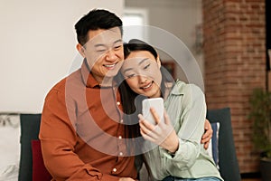 Loving happy korean spouses video calling via cellphone, middle aged man and his young wife relaxing on sofa at home