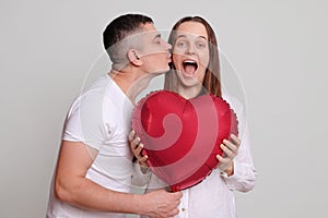 Loving husband kissing amazed excited wife on Valentines Day celebrating together Caucasian man and woman holding heart shaped air
