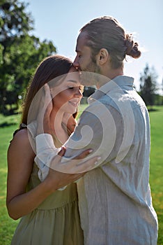 Loving husband kissing smiling young wife in forehead while they posing together outdoors in summer park