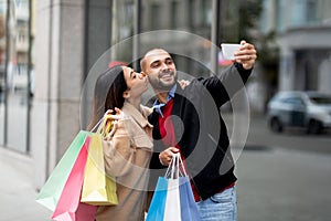 Loving interracial couple with colorful bags taking selfie on mobile phone in front of shopping mall