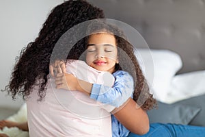 Loving little black girl in casual wear hugging her mother on bed at home. Strong family relationships concept photo
