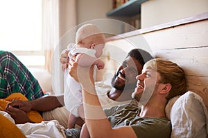 Loving Male Same Sex Couple Cuddling Baby Daughter In Bedroom At Home Together