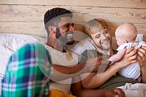 Loving Male Same Sex Couple Cuddling Baby Daughter In Bedroom At Home Together