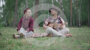 Loving man in hat playing ukulele for girlfriend or wife in summer park as blurred dog passing camera at front. Portrait