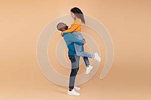 Loving mature black man lifting his wife and holding her on peach studio background, profile side view, free space