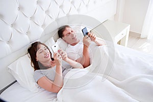 Loving middle aged couple smiling and watching something on her modile phones in bedroom, happy people