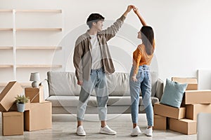 Loving millennial Asian couple dancing in their new home among carton boxes on moving day, copy space