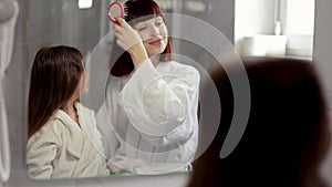 Loving mom brushing hair of cute little girl in the bathroom.