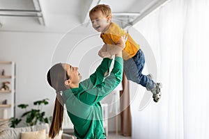 Loving mom lifting her little son in air while spending time together at home, holding kid in hands and smiling