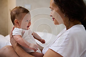 Loving mom sitting on the bed, holding her baby boy, communicating and smiling to him, establishing emotional contact