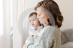 Loving mother kissing cute baby near window at home