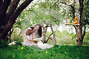 loving mother reading book to toddler son outdoor on picnic in spring or summer park