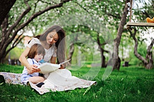 Loving mother reading book to toddler son outdoor on picnic in spring or summer park. Happy family and mothers day