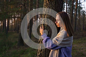 Loving nature concept: relaxed woman hugging tree trunk in autumn forest with closed eyes at sunset