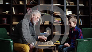 Loving old senior grandfather and grandson are playing chess sitting at desk in cozy living room.