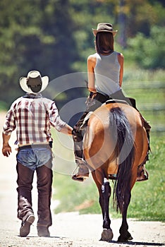 Loving the outdoors. Rear view of a cowboy leading a young woman on a horse along a country lane.