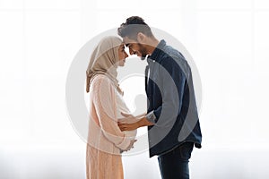 Loving pregnant muslim couple bonding together at home, standing near window