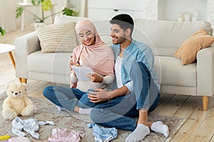 Loving pregnant muslim couple getting ready for childbirth, making checklist while sitting with baby clothes around