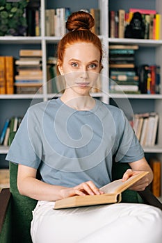 Loving redhead young woman student is reading book enjoys of rest at home office, looking at camera.